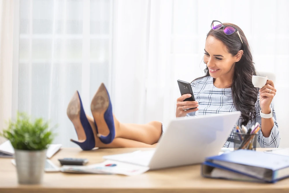 mujer procrastinando oficina pies sobre mesa tomando cafe sonriendo su telefono celular